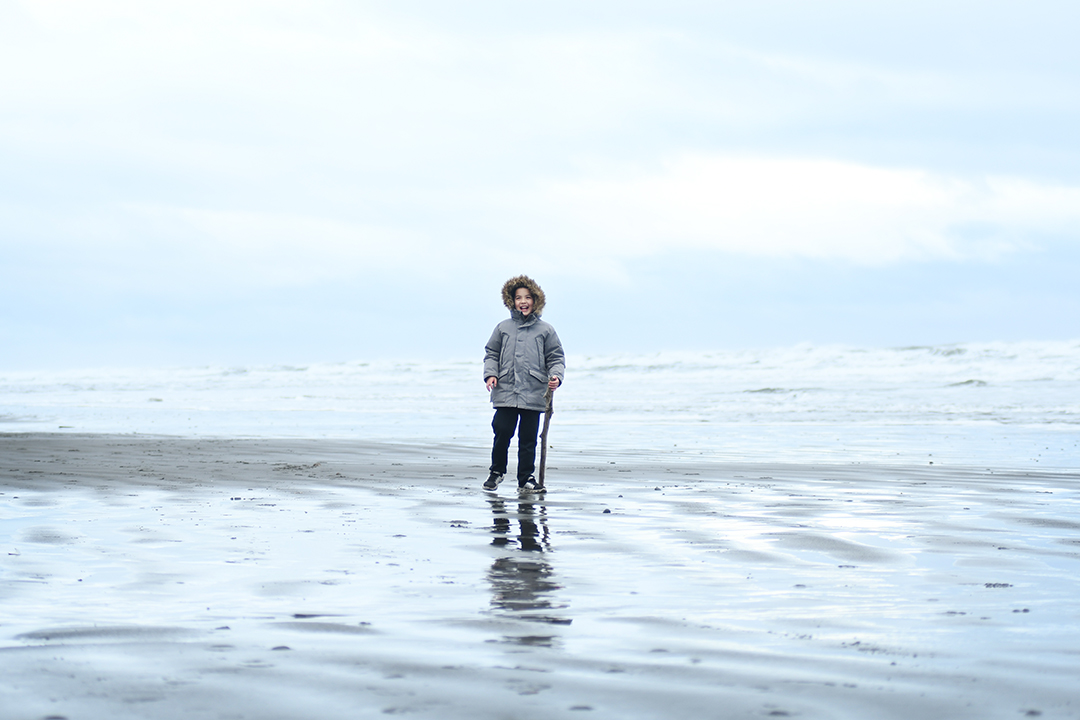 Boy in winter jacket with furry hood standing along on a wide expanse of Pacific Ocean beach with glass reflection from the water