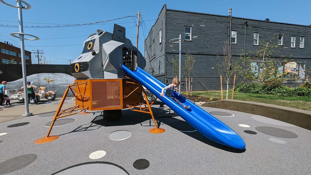A preschool age boy descends a blue slide coming from a replica lunar landing play structure part of the playground at Kent's updated Kherson Park