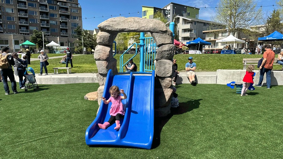 Young girl slides down a small blue slide at the new Ballard Commons Park playground in Seattle.
