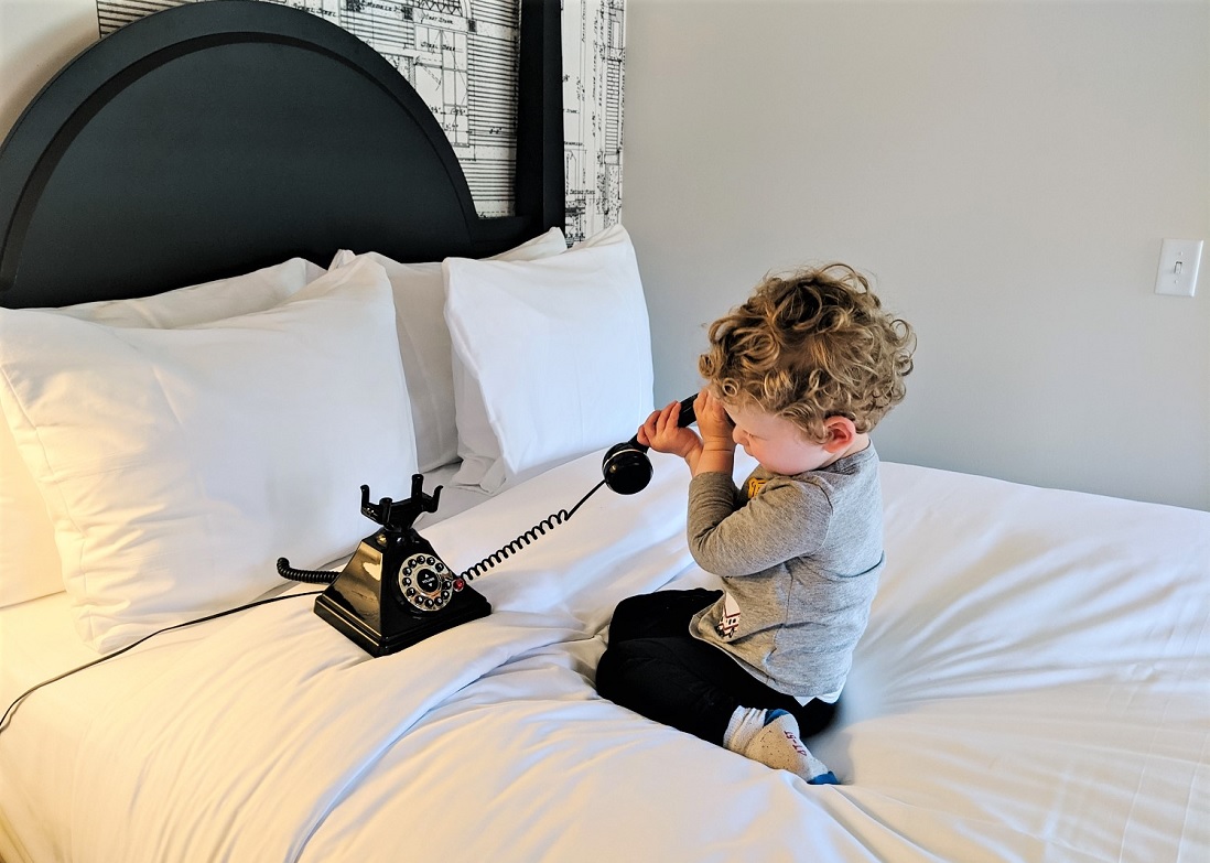 Young boy sitting on a hotel bed playing with an antique rotary dial phone at new Lodge at St. Edward State Park near Seattle