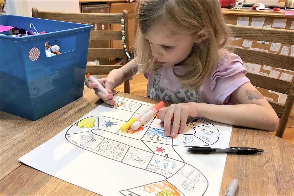 Young girl sitting at a table with markers and paper creating a homemade board game virtual travel activities