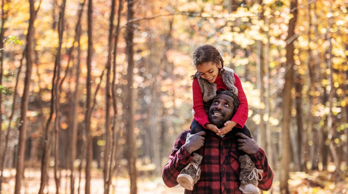 father with daughter on his shoulders surrounded by trees with fall colors