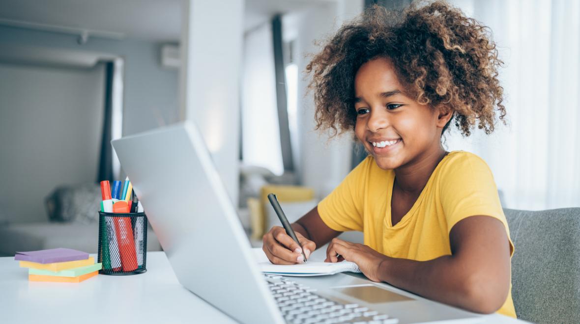black girl in a yellow tee shirt on a computer smiling with school supplies on the table next to her