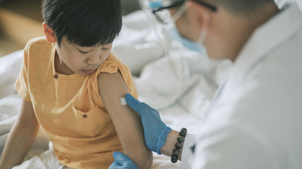 small boy looking down at his arm as a masked doctor puts a bandaid over a vaccine shot poke
