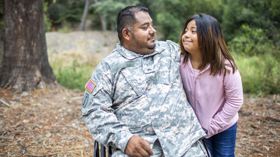 Veteran dad in army fatigues and sitting in wheelchair smiles at and hugs daughter of about age 11. she is smiling at him, wearing a pink sweatshirt