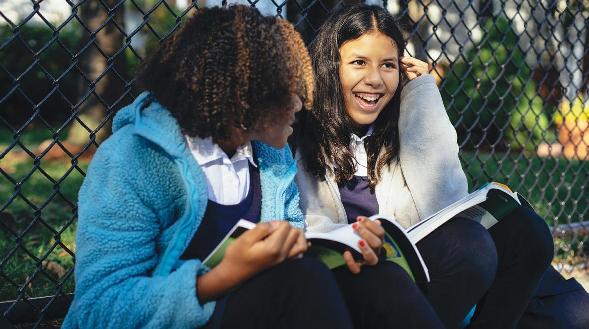 Two girls sitting by a fence smiling and holding school books 