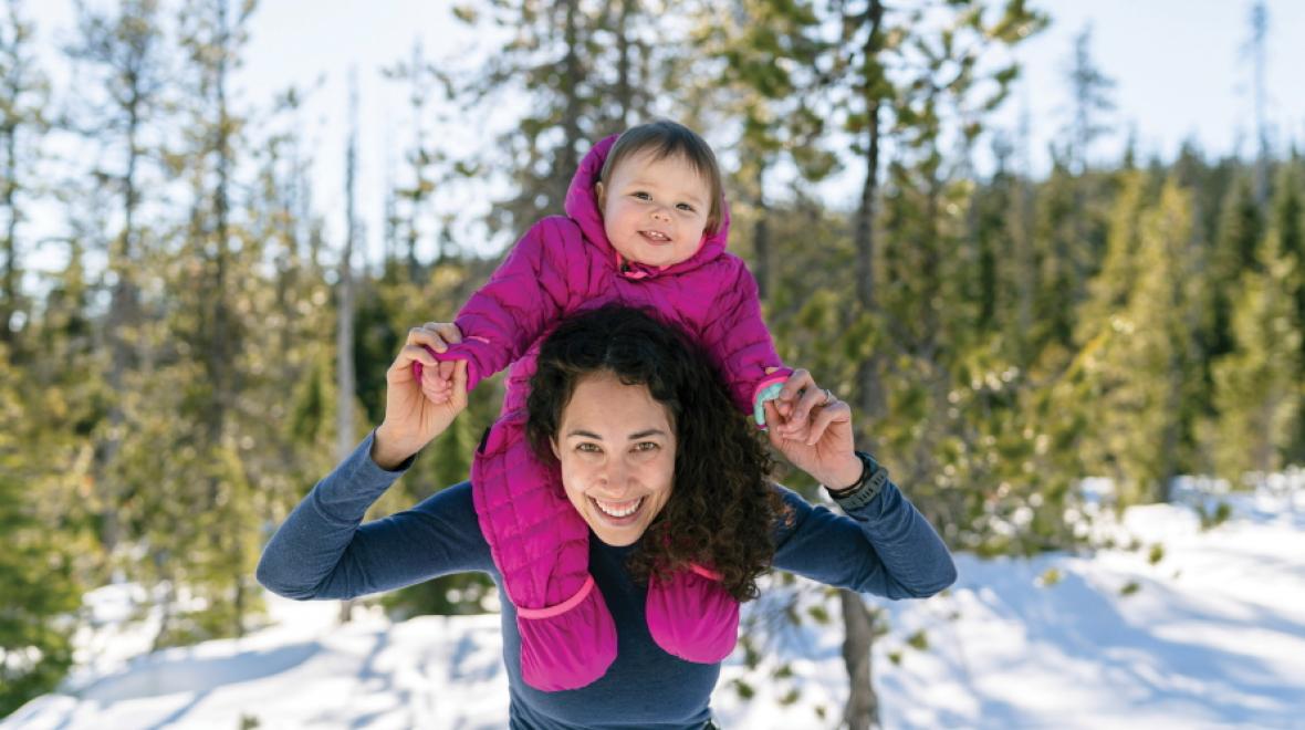Woman holding a smiling baby on her shoulders in the woods with snow on the ground