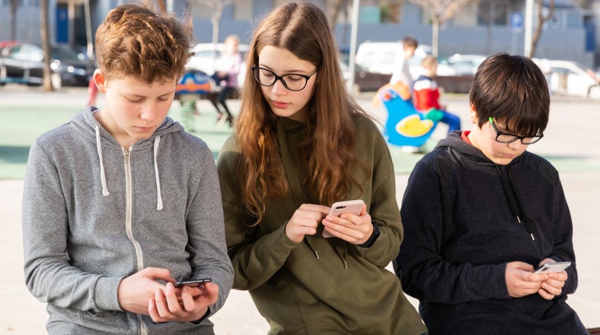 Three tweens in hoodie sweatshirts look at their phones while leaning on a park bench best tween and teen outings around Seattle
