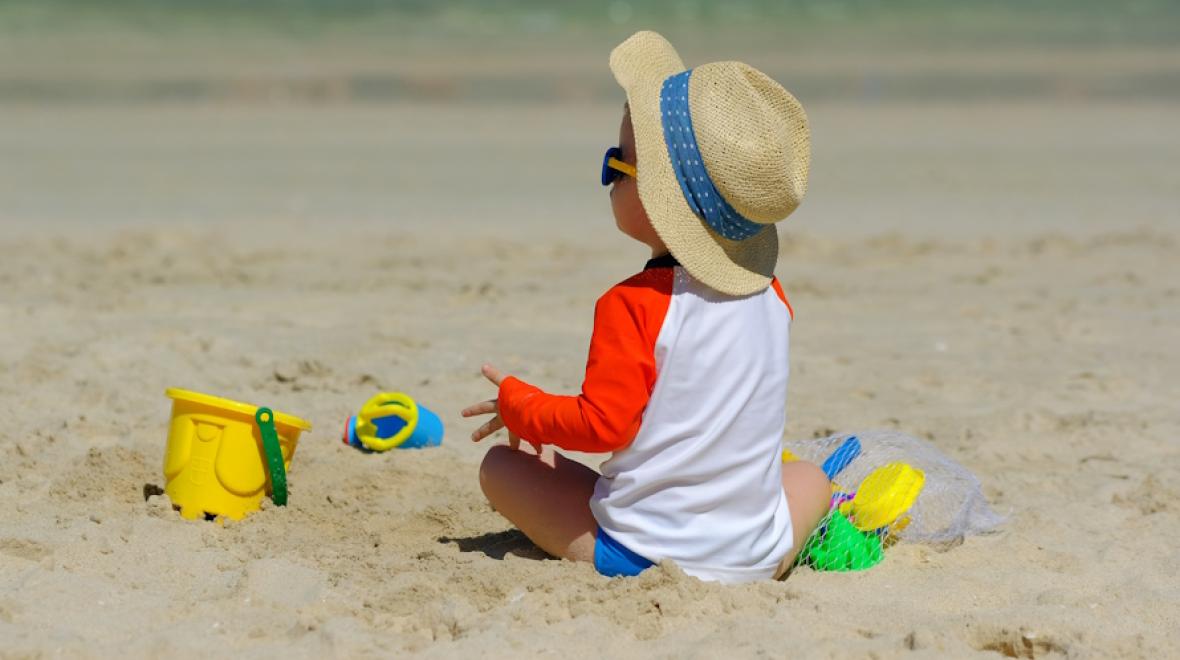 Toddler sitting on the beach wearing sun protection hat, sunglasses and rash guard