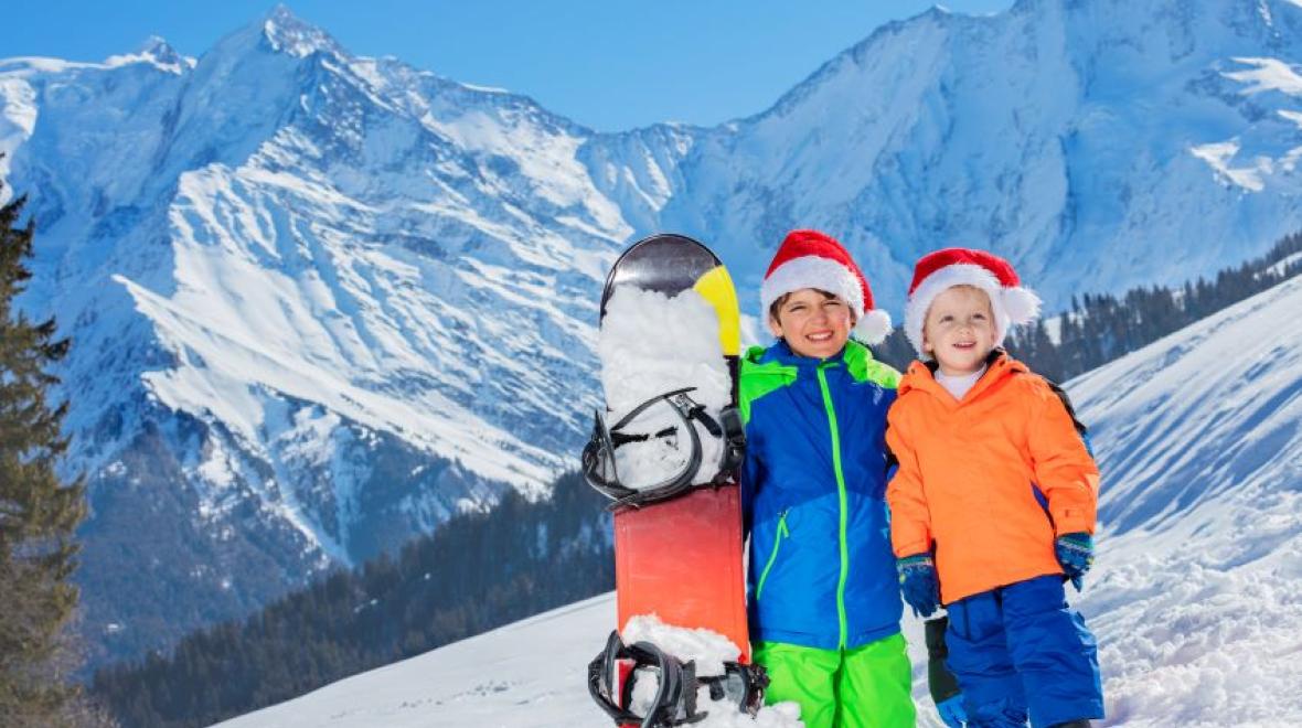 Two happy boys in Santa hats stand with snowboard over the mountains on background on Christmas Day