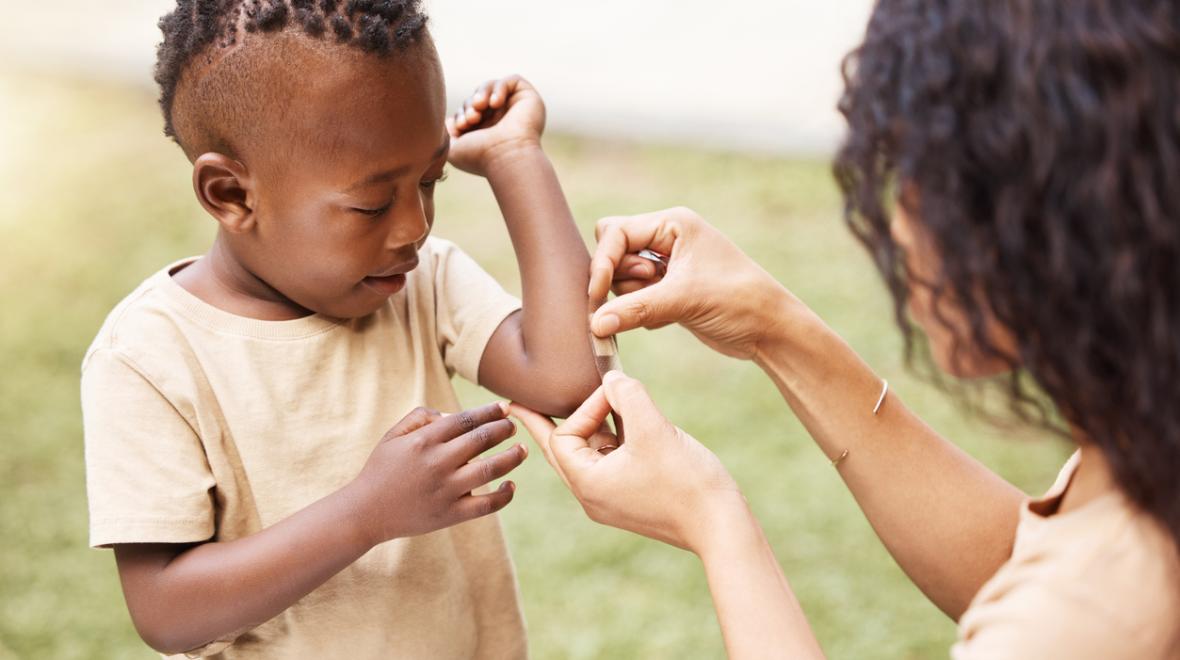 A young child gets a bandaid gently placed on his elbow by a caregiver.