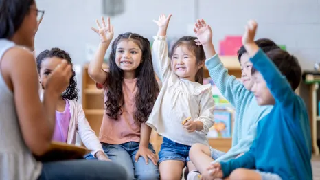 a classroom of preschool children raise hands to answer a question from the teacher