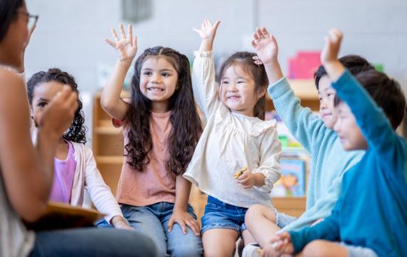 a classroom of preschool children raise hands to answer a question from the teacher