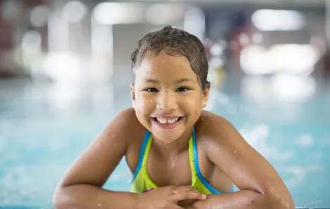 Elementary-age girl smiling, wearing swimsuit, with arms resting on the edge of an indoor swimming pool
