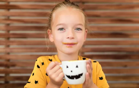 girl in an orange halloween shirt drinking out of a white jack o'lantern mug