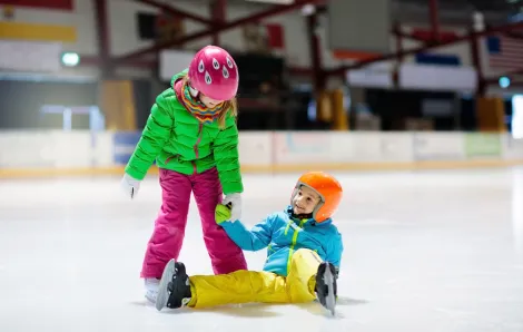 Kids in colorful winter clothes and wearing helmets ice skate at an indoor ice rink one has fallen and the other is helping her up