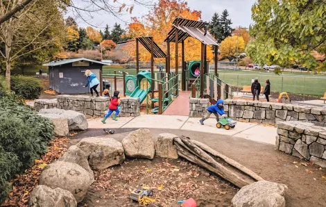 Kids play at updated Lakewood Playground in South Seattle. There's a sand pit in the foreground and a new play structure in the background and fall foliage viewable as well