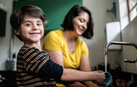Boy washing a mug and smiling at the camera with mom in the background