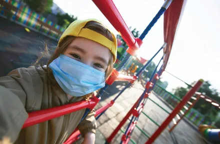 closeup of a girl in a mask holding onto the bars of a metal play structure