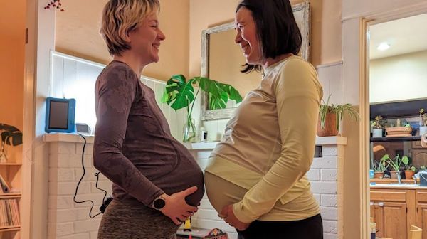 Pregnant mom friends smile at each other and touch bellies.