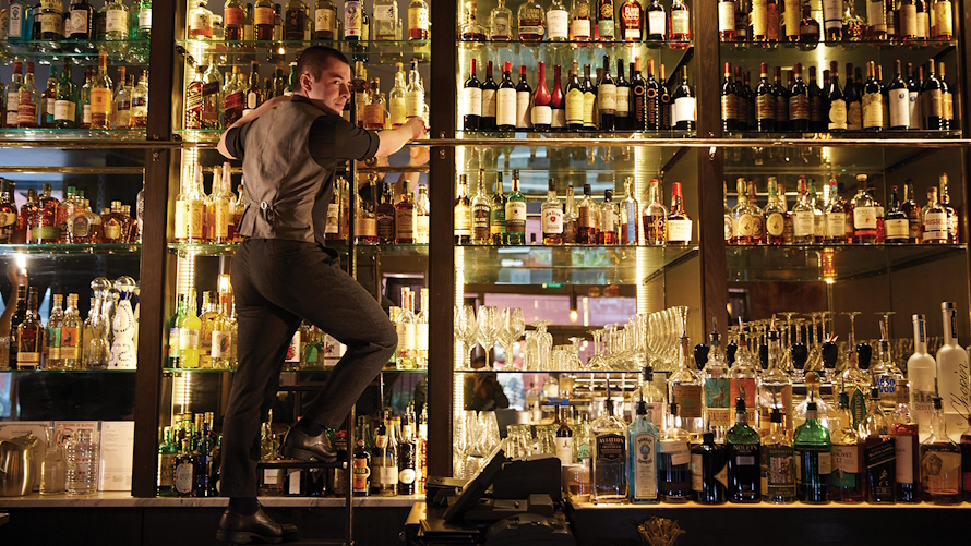 "A man on a ladder in front of a large wall of liquor bottles at Central Bar + Restaurant"