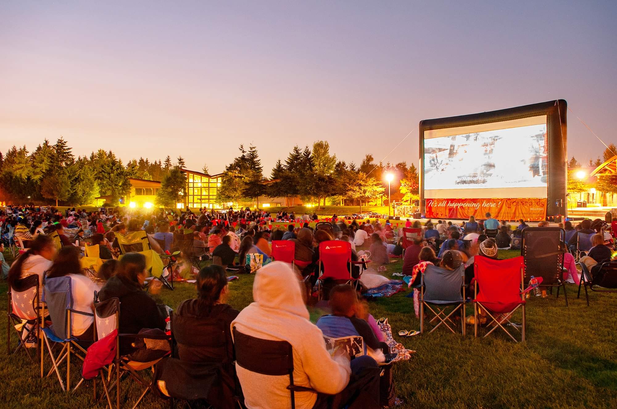A view of people sitting on the lawn at Crossroads Park in Bellevue watching a movie outdoors on a big screen on a summer evening
