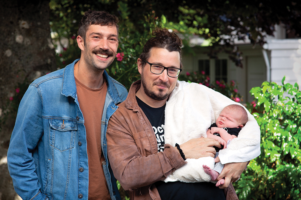 "Two dad's smiling and holding their newborn son"