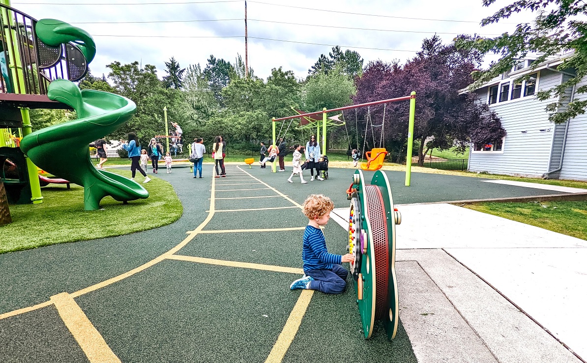 A boy plays with a sensory panel game with swings and a merry-go-round climber in the background at the new Train Park playground at Mercer Island’s Mercerdale Park