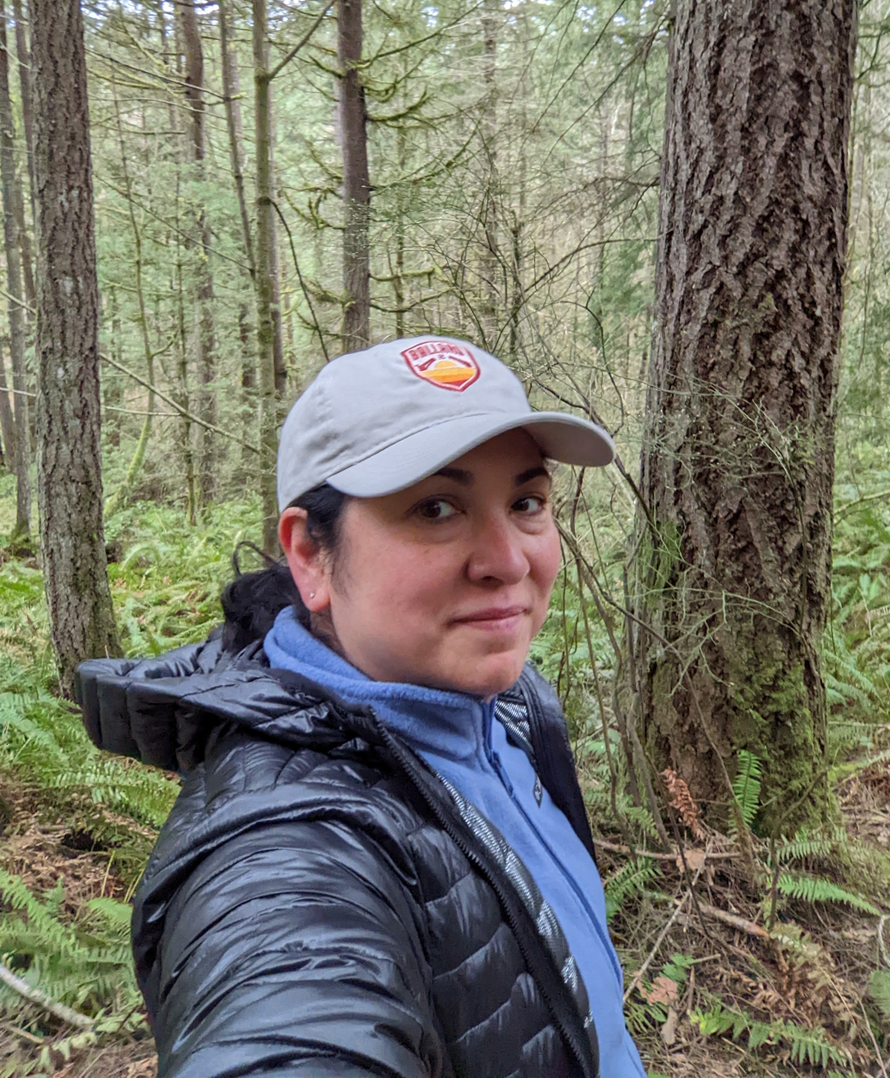 "Woman standing in the forest wearing a black puffy coat and baseball hat"