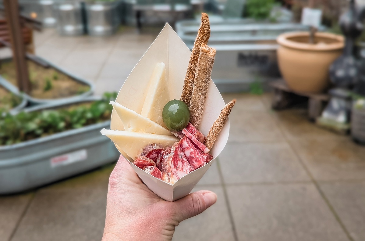 View of a hand holding a paper cone with charcuterie snacks inside from DeLaurenti’s find foods including salamai, breadsticks, olives Pike Place Market best snacks with kids