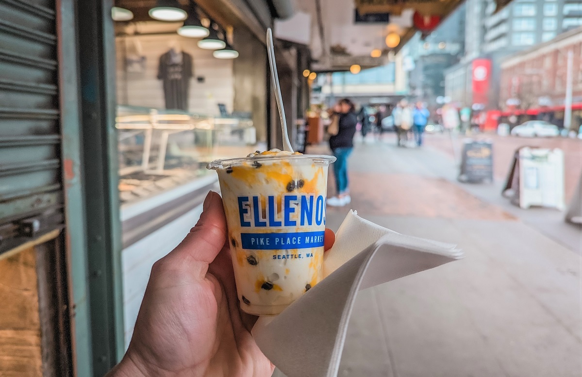View of a cup of Ellenos yogurt from Pike Place Market being held in a hand and enjoyed as a walking snack during a family market day