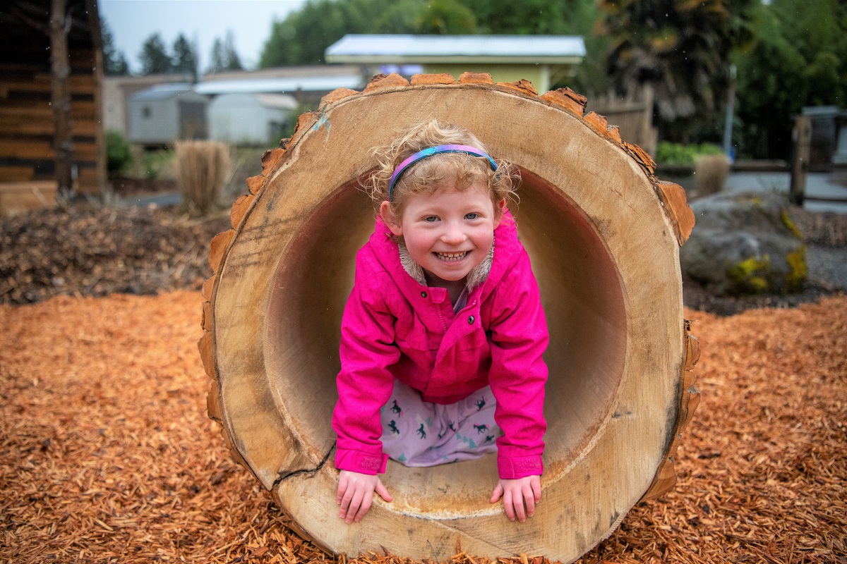 A smiling young girl in a pink jacket looks out from a hollow log she’s playing in at PDZA’s new Little Explorers Nature Play Garden opening spring 2022