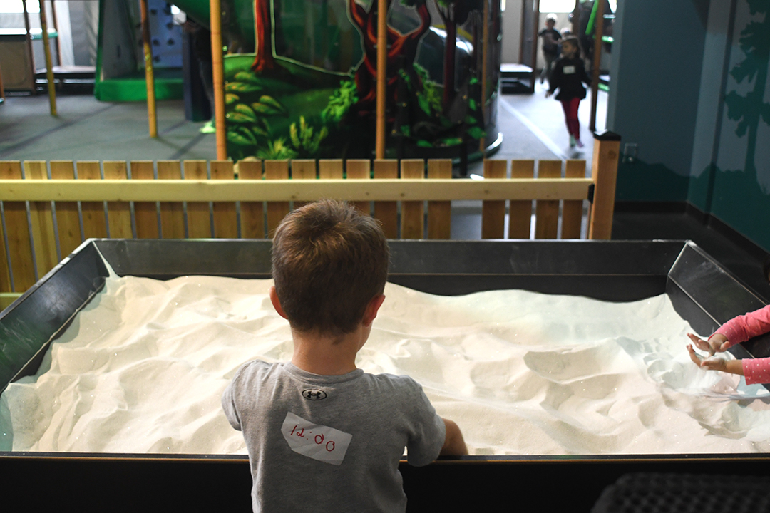 A young boy with a sticker on his back showing his exit time from The Ridge Activity Center in Bothell plays at the sand table among other great indoor play options