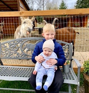 An 8-year-old blond boy in a blue jacket holds a baby on his lap while a goat sticks his tongue out behind them at Sammamish Animal Sanctuary