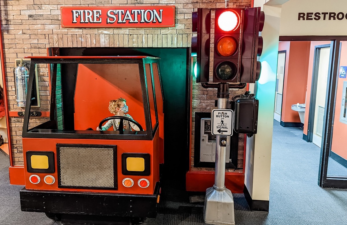 Young boy with curly hair wearing face mask sits in the driver's seat of a life-size toy fire truck at the reopened Seattle Children's Museum at Seattle Center