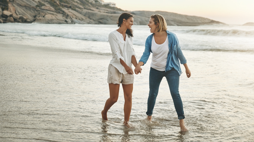 "Two women holding hand and laughing on the beach on a kid-free vacation"