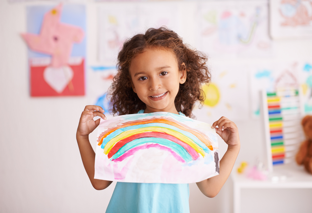 "Child smiling and holding up painting of a rainbow"