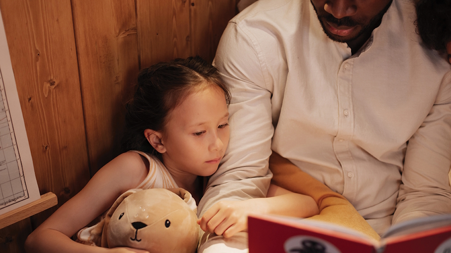 "Young girl holding a stuffed animal sitting close to her dad who is reading her a book""