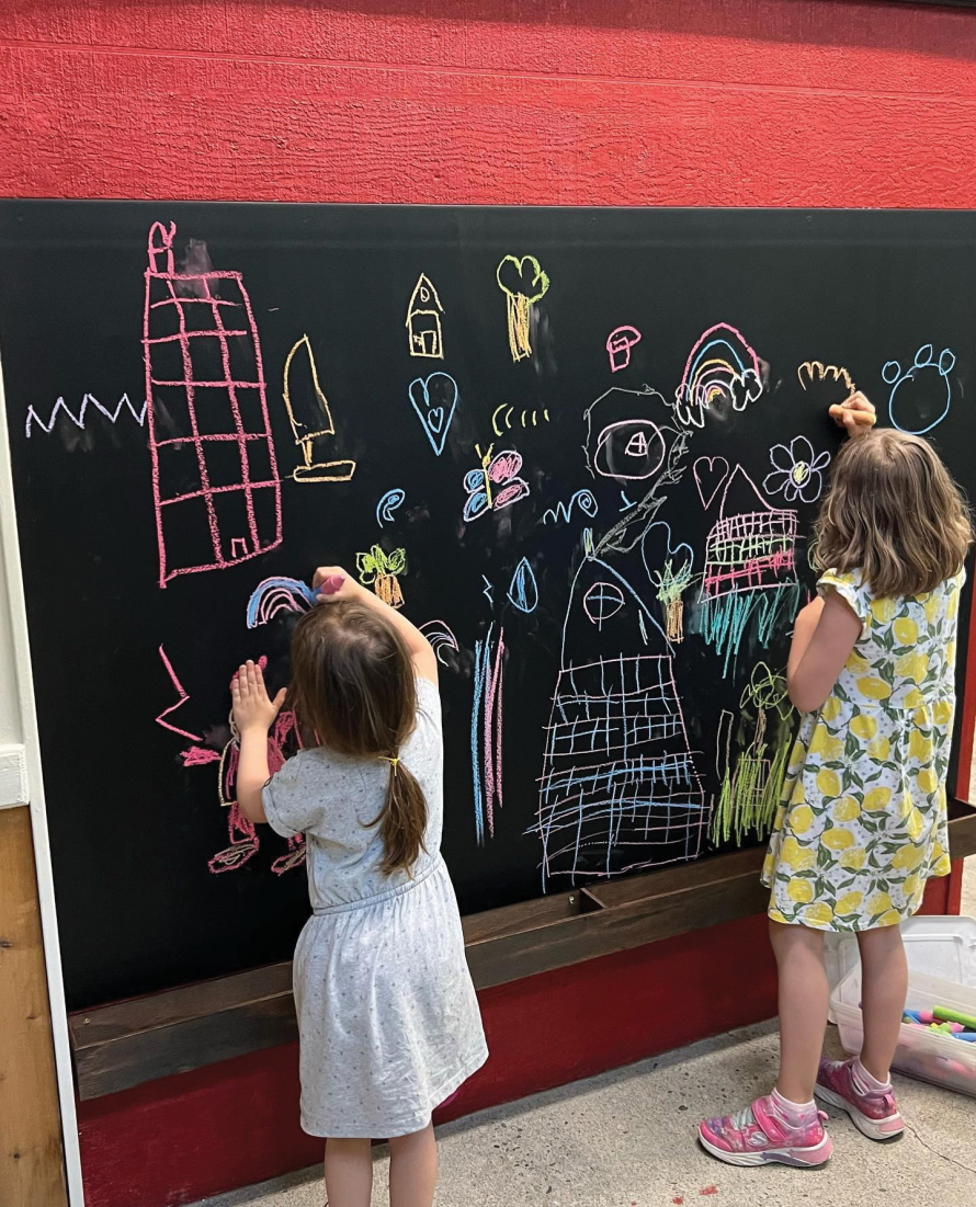 "Two young girls drawing with chalk on a blackboard wall"