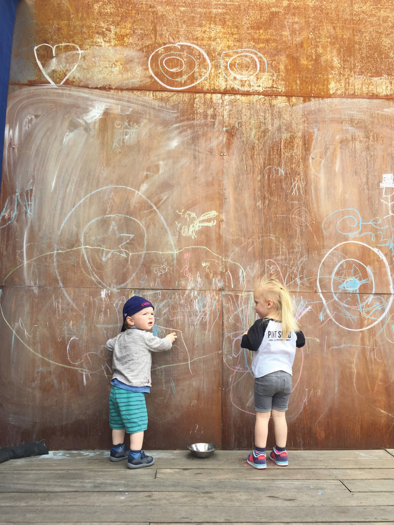 "Two young children standing next to a large wall drawing on it with chalk"