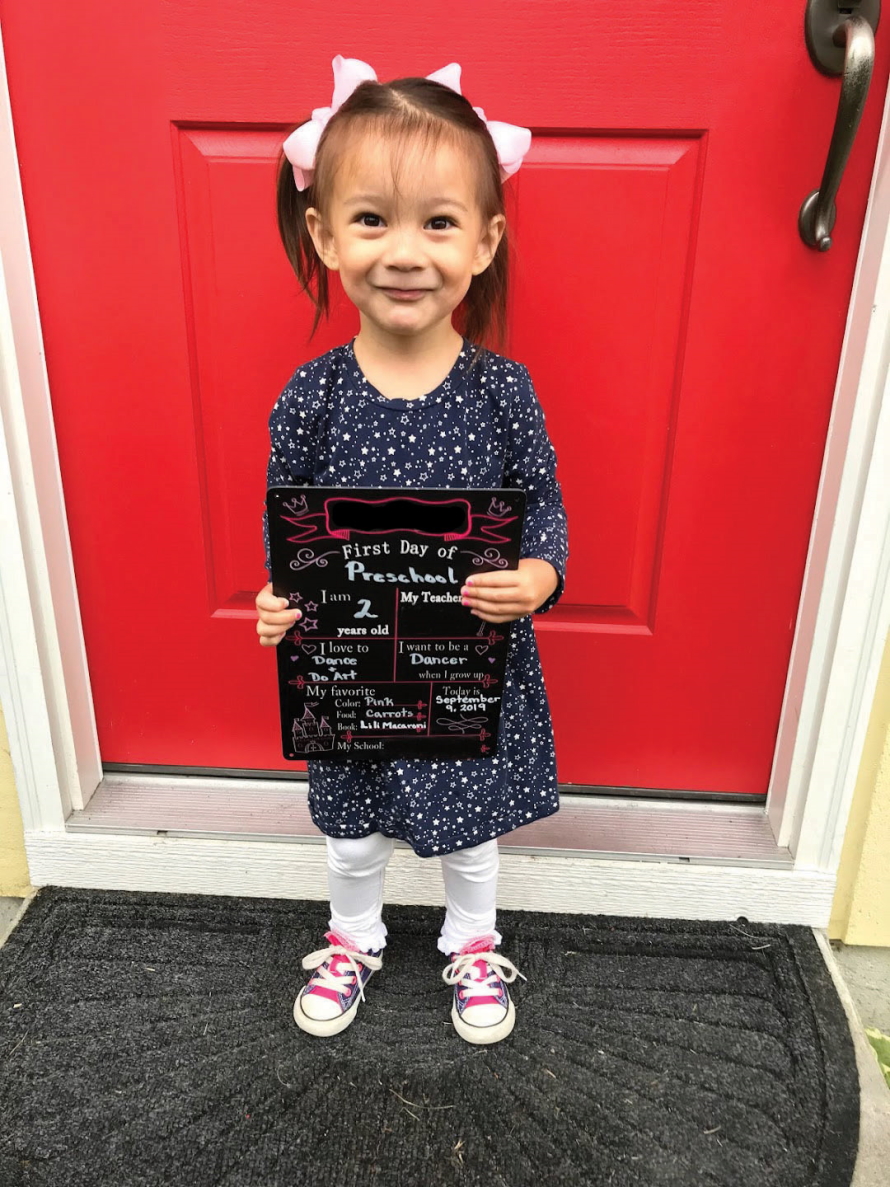 "Little girl standing in front of a red door on the first day of school"