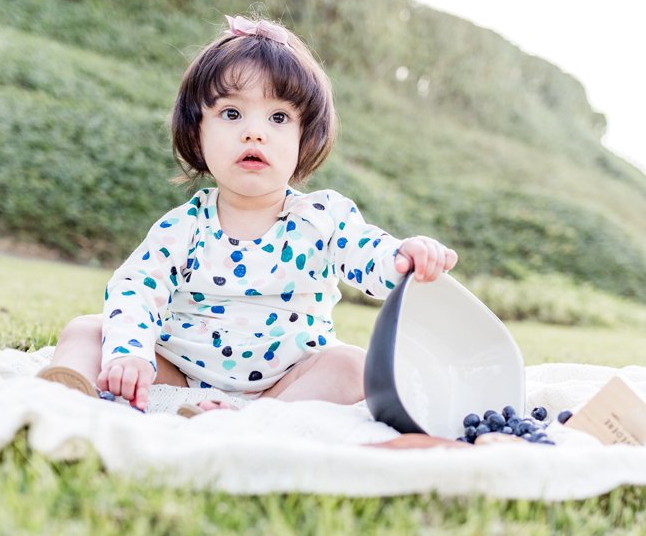 "Baby sitting on a blanket in a field with a bowl of spilling blueberries"