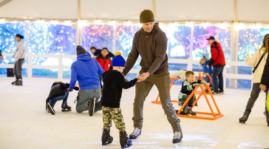 A father and his young son skate on the ice at a seasonal rink.