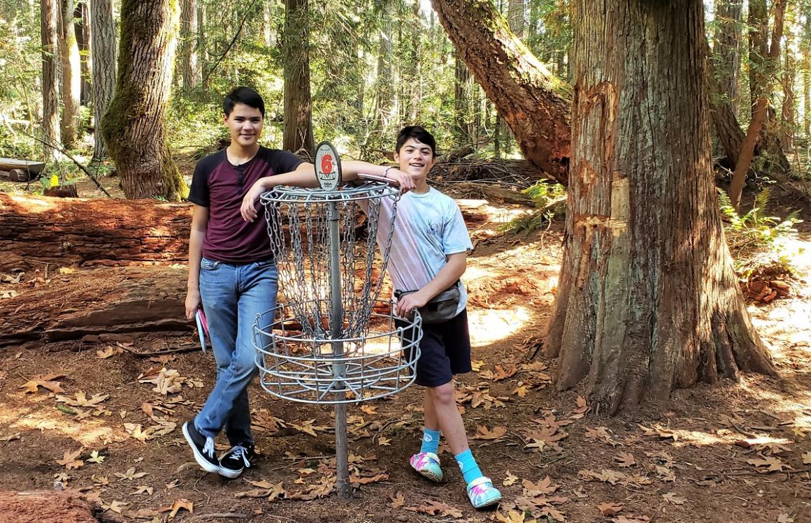 Two teen boys pose with a disc golf basket-hole at a disc golf course near Seattle where to try this fun and free family sport