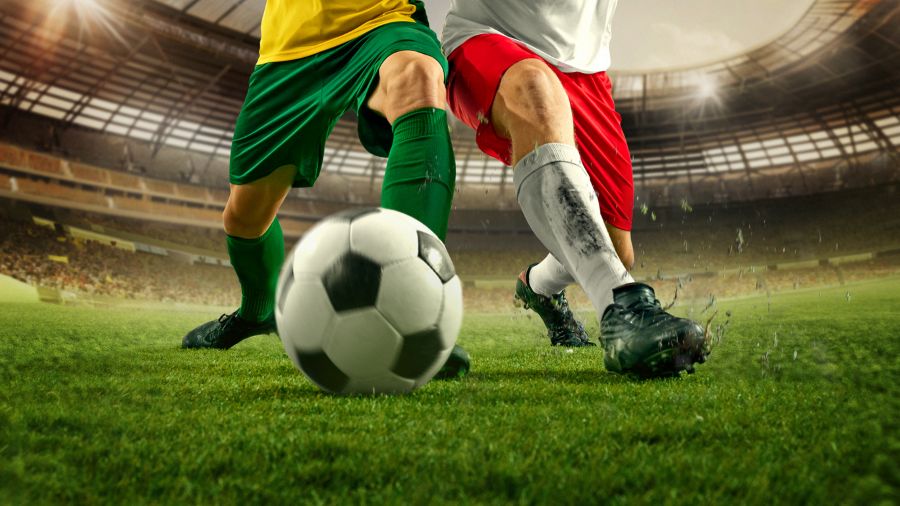 Two soccer player vie for the ball as they both try to kick it on a grassy field.