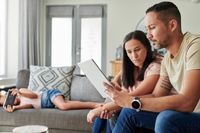 Parents on coach reading tablet with child in background