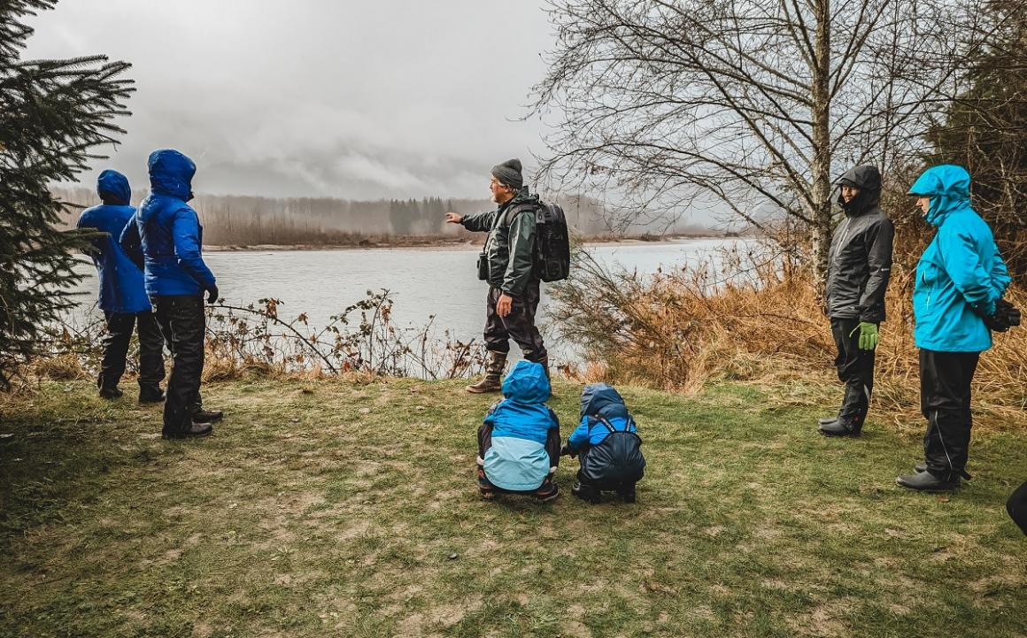 Adults and kids stand near the bank of the Skagit River on a rainy winter day listening to educational tour given by Joe Ordonez of Skagit River Bald Eagle Intepretive Center