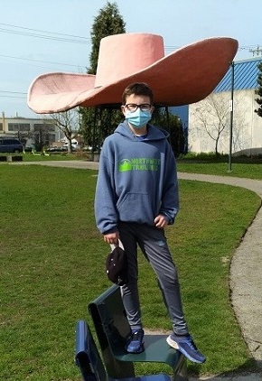 Boy age 12 standing in front of the hat of Hat and Boots scultpure at Seattle’s Oxbow Park positioned to appear as though the hat is on his head