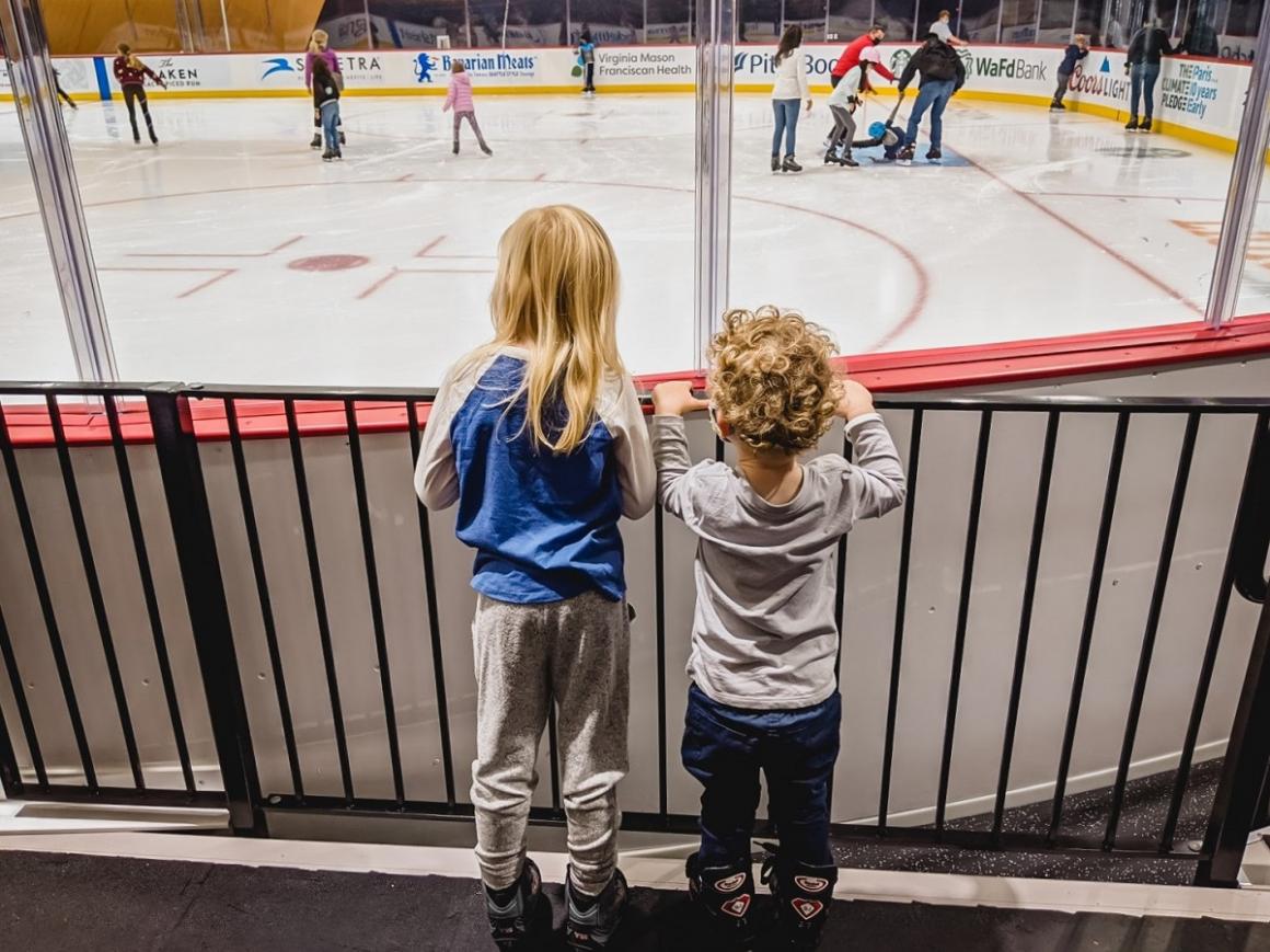 Two young kids in brand-new hockey-style rental skates look out onto the ice at the new Kraken Community Iceplex in Seattle