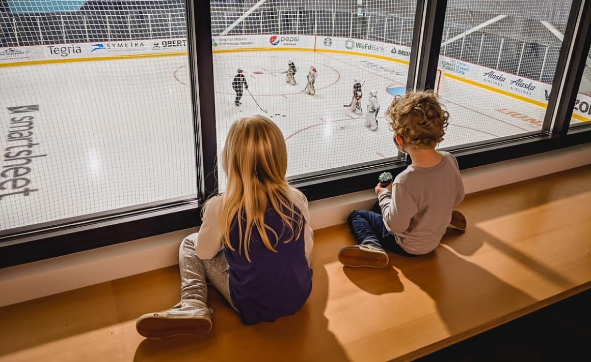 Kids sitting on a windowside bench look out over the ice where a girls' hockey team practices at the new kraken community iceplex in seattle